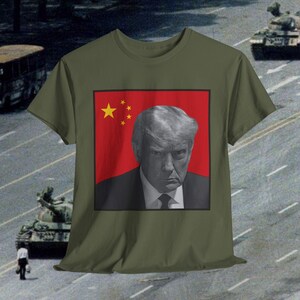 May include: Olive green t-shirt featuring a graphic of a stern-faced man against a red background with yellow stars, reminiscent of a flag. The image is set against a backdrop of tanks and a street scene.