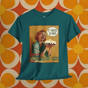 Puede incluir: Camiseta verde azulado con un gráfico retro de una mujer sosteniendo un bizcocho de limón. El gráfico incluye el texto "It tastes so nice!" en un bocadillo y "Mama's Lemon Pound Cake" debajo.