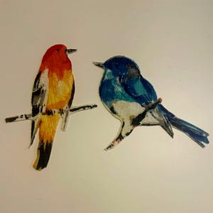 May include: Two bird-shaped cutouts featuring painted textures. The left bird has a bright orange head and chest with yellow and grey wing details, perched on a branch. The right bird is blue with a white belly and dark grey wing markings.