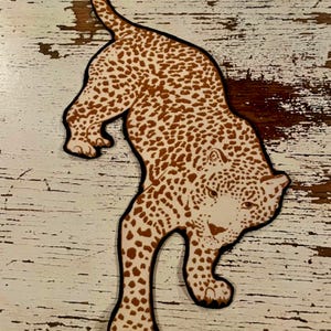May include: A flat, die-cut cutout of a leopard in a stalking pose. The leopard features a cream-colored base with brown spots and a distinct black outline. The animal is depicted with its head lowered and tail curved upward in a prowling motion.