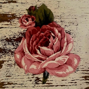 May include: A decorative floral decal featuring a large, multi-layered pink rose with white highlights and a smaller rosebud with dark green leaves. The design is set against a distressed, cream-colored background with visible brown wood grain.