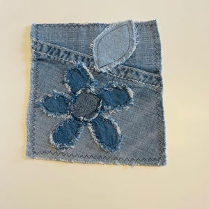 May include: A denim patch with a blue flower and leaf appliqué. The flower has a frayed edge, and the patch is stitched with dark blue thread. The background is a light blue denim.