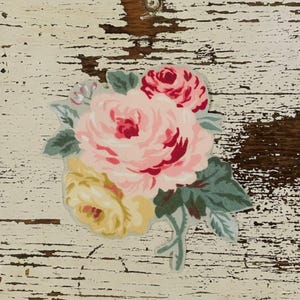 Op de afbeelding: Een bloemensticker met een boeket rozen in roze, rode en gele tinten, met groene bladeren. De sticker is geplaatst tegen een verweerde witte en bruine houten achtergrond.