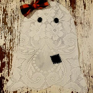 May include: A white lace ghost decoration featuring two black circular button eyes, a small black square fabric patch with white stitching, and a red and black buffalo plaid fabric bow attached to the top corner of the lace material.