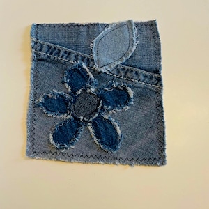 May include: A square denim patch featuring a pocket design with a frayed-edge dark blue denim flower appliqué and a single leaf appliqué. The edges of the light blue denim square are finished with a black zigzag stitch, creating a textured look.