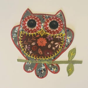 Pode incluir: Aplique de tecido em forma de coruja, com um padrão floral no corpo e um exterior com padrão cinza e vermelho. A coruja tem olhos de botão pretos e está pousada num galho verde com folhas. Costuras amarelas delineiam o desenho.
