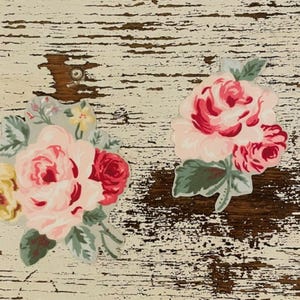 Op de afbeelding: Twee bloemenuitsparingen met roze, rode en gele rozen met groene bladeren. De bloemen zijn geplaatst tegen een verweerde, gebroken witte en bruine houten achtergrond, wat een vintage esthetiek creëert.