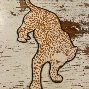 Può includere: Ritaglio di legno a forma di leopardo con un motivo maculato marrone e contorno nero. Il leopardo è raffigurato in posizione di camminata, con la coda ricurva verso l'alto. Il ritaglio è posto su una superficie di legno bianco invecchiato.