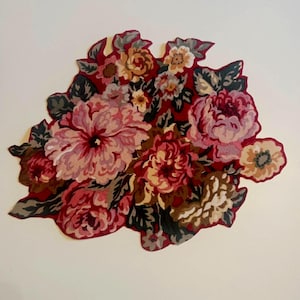 May include: A tufted rug shaped like a floral bouquet featuring large, layered blossoms in shades of deep red, soft pink, and muted cream. The design includes dark green and grey foliage accents, with a solid red border outlining the organic shape.