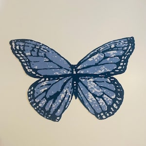 May include: A blue butterfly illustration with dark blue outlines and white accents. The butterfly's wings are spread wide, showcasing a detailed pattern. The artwork is set against a cream-colored background, creating a striking contrast.