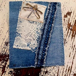 May include: A denim patch with a dragonfly embroidery on a white square, a white lace section, and a frayed denim strip. The patch is stitched onto a larger denim square, creating a textured, handcrafted look.