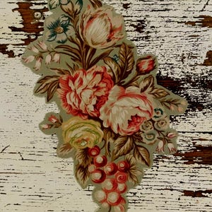 May include: A die-cut paper cutout featuring a floral arrangement with large pink and cream roses, smaller white blossoms, and clusters of red berries. The design is set against a muted sage green background with detailed brown foliage and stems.