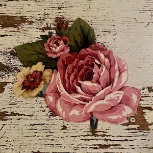 May include: A floral decal featuring a large, layered pink rose as the central element, accompanied by a smaller pink rosebud, a pale yellow flower with a dark red center, and several dark green leaves, all arranged in a cluster on a white surface.