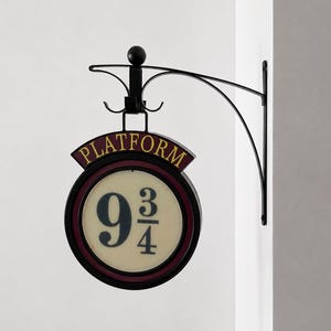 May include: A circular wall-mounted sign featuring the text Platform 9 3/4 in black lettering on a cream background with a dark red border. The sign is held by a black metal bracket with a decorative curved arm and a small sphere finial on top.