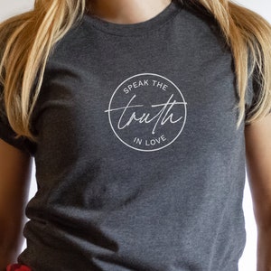 May include: Dark gray t-shirt with the text "Speak the Truth in Love" in a white circular design. The person is wearing a red and black plaid shirt tied around the waist.