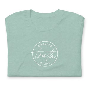 May include: A folded, light green t-shirt with a white circular design. The design features the words "Speak the truth in love" in a mix of script and block lettering.