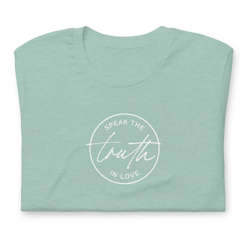 May include: A folded, light green t-shirt with a white circular design. The design features the words "Speak the truth in love" in a mix of script and block lettering.