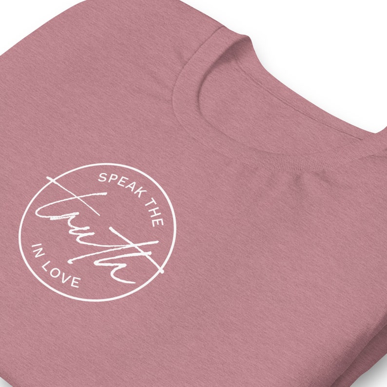 May include: Dusty rose-colored t-shirt with a white circular graphic. The graphic features the words "Speak the Truth in Love" in a handwritten font, encircled by a white line. The t-shirt is folded, showing the neckline and a portion of the front.