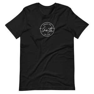 May include: Black t-shirt with a white circular design. The design features the words "Speak the Truth in Love" in a handwritten font. The t-shirt is made of a soft material and has short sleeves and a crew neck.
