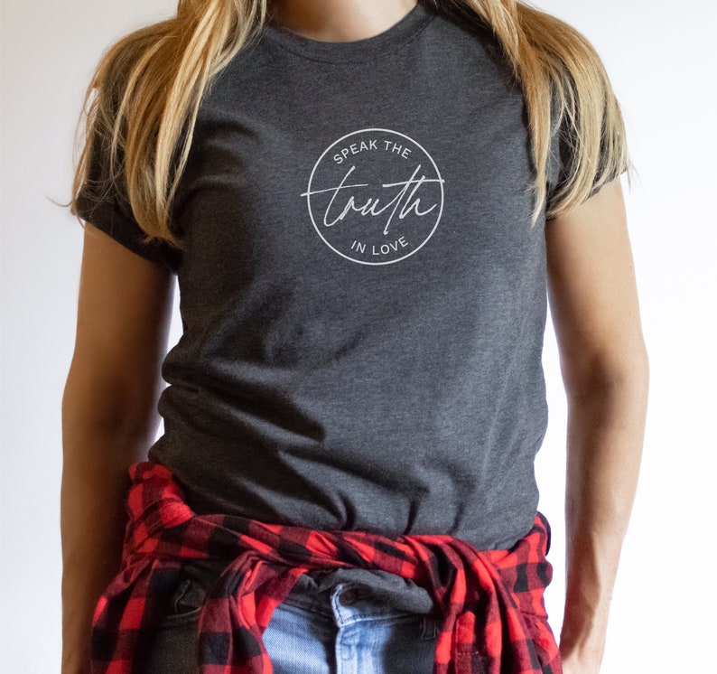 May include: Dark gray t-shirt with the text "Speak the Truth in Love" in a white circular design. The person is wearing a red and black plaid shirt tied around the waist.
