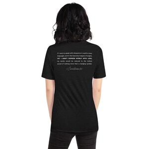 May include: Black t-shirt with white text on the back. The text includes a quote from 1 Corinthians 13:1 about the importance of love. The shirt is a short-sleeved, unisex style.