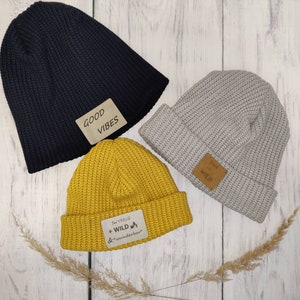 Könnte beinhalten: Drei Strickmützen in verschiedenen Farben mit Patches. Die schwarze Mütze hat einen braunen Patch mit der Aufschrift "GOOD VIBES". Die graue Mütze hat einen braunen Patch mit der Aufschrift "fresh + WILD". Die gelbe Mütze hat einen braunen Patch mit der Aufschrift "Sei Frech + WILD & wunderbar".