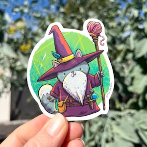 May include: A gray cat wearing a purple wizard robe and a pointed hat with a yellow band. The cat is holding a staff with a pink ball of yarn on top. The cat has a white beard and a small pouch on its belt. The background is a green circle with a geometric pattern.