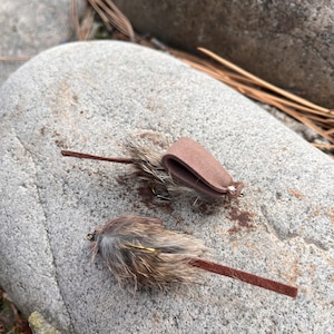 May include: Two fishing lures, designed to resemble small rodents, rest on a gray stone. Each lure features a brown suede tail and a body of mottled brown and gray fur. One lure has a brown leather loop.