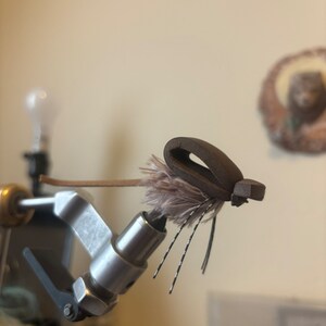 May include: A brown and grey fly fishing lure with feathers and a brown leather body is secured in a vice.