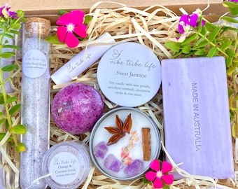 Puede incluir: Una caja de regalo llena de productos de baño y cuerpo con temática de lavanda. La caja incluye una vela con aroma a lavanda, una barra de jabón, un tubo de exfoliante corporal de café, un tarro de exfoliante de naranja y coco, y una bomba de baño de lavanda. La caja está decorada con flores y paja.