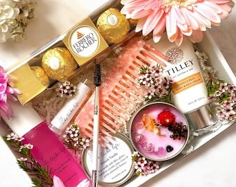 May include: A gift box containing a box of Ferrero Rocher chocolates, a pink hair comb, a Tilley hand and nail cream tube, a lip balm, a spoolie brush, a pink candle with crystals and dried flowers, and a pink spray bottle, decorated with pink flowers.