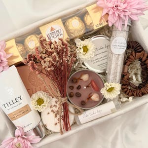 May include: A gift box containing a Tilley vanilla bean hand and nail cream, Ferrero Rocher chocolates, a Vibe Tribe Life coffee body scrub, a soy candle with coffee beans and shells, a lip balm, a brown scrunchie, dried flowers, and pink flowers.