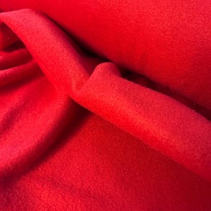 May include: A close-up of a vibrant red fleece fabric. The material is soft and textured, with visible fibers. The fabric is folded and rolled, showcasing its drape and volume. Ideal for apparel and craft projects.