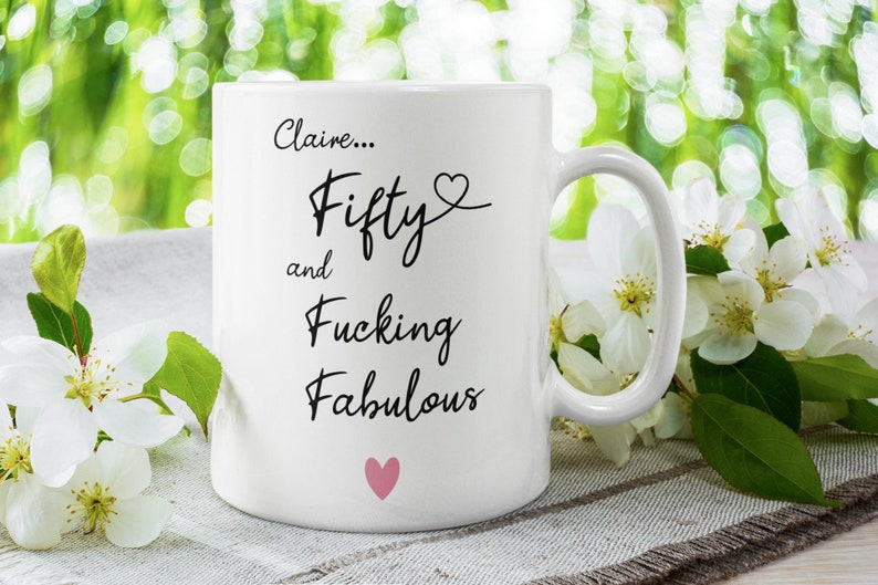May include: White ceramic mug with black text that reads "Claire... Fifty and Fucking Fabulous" with a pink heart above the word "Fifty" and a pink heart below the word "Fabulous".