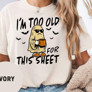 May include: An ivory t-shirt featuring a graphic of a ghost wearing sunglasses and holding a brown mug. The text reads I'M TOO OLD FOR THIS SHEET. Small black bats are scattered around the ghost, and the shirt has a crew neckline and short sleeves.