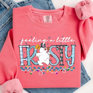 Puede incluir: Una sudadera de color coral con la frase "feeling a little FROSTY" y un gráfico de un muñeco de nieve. La palabra FROSTY está en letras a cuadros azules y blancos. Una cadena de luces de colores está en la parte inferior.