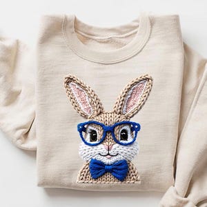May include: A beige sweatshirt featuring an embroidered bunny wearing blue glasses and a blue bow tie. The bunny has knitted ears with pink and white details. The sweatshirt has long sleeves with rolled-up cuffs.