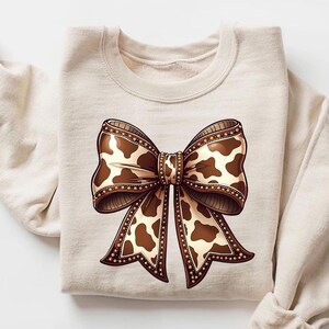 Graphic sweatshirt featuring an illustrated ribbon bow with cow print pattern inside the loops and tails; centered decorative bow design with rounded loops and flowing ribbon ends; western-inspired cow pattern with stitched border detailing; symmetrical layout across the front of the sweatshirt; clean illustrated artwork designed for casual everyday wear; professionally printed graphic with sharp outlines and clear pattern detail.
