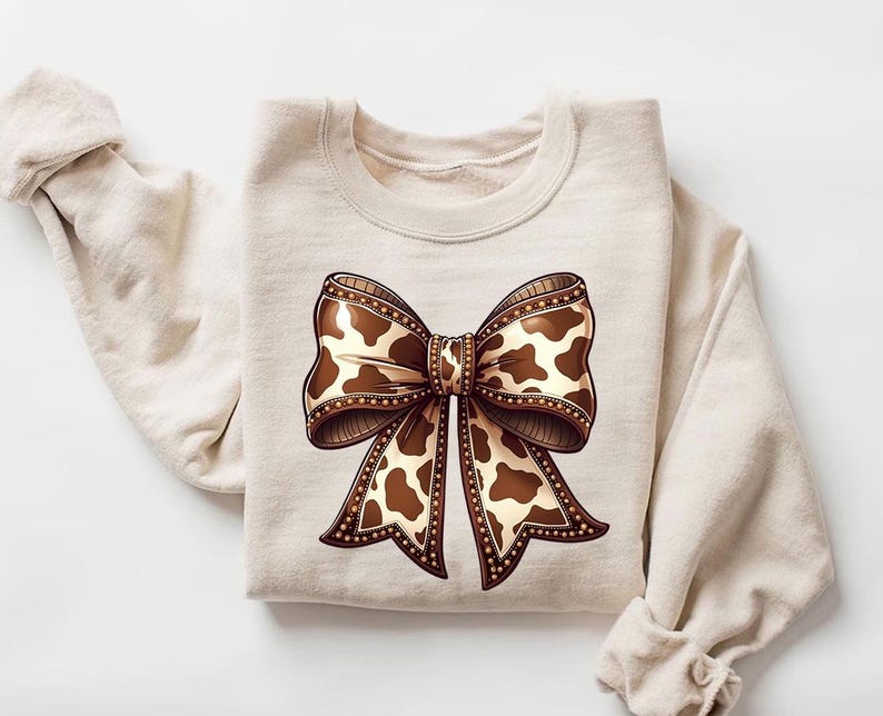 Graphic sweatshirt featuring an illustrated ribbon bow with cow print pattern inside the loops and tails; centered decorative bow design with rounded loops and flowing ribbon ends; western-inspired cow pattern with stitched border detailing; symmetrical layout across the front of the sweatshirt; clean illustrated artwork designed for casual everyday wear; professionally printed graphic with sharp outlines and clear pattern detail.