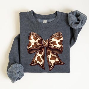 May include: A dark gray sweatshirt featuring a large bow design. The bow has a brown and white cow print pattern with a decorative border. The sleeves are rolled up.