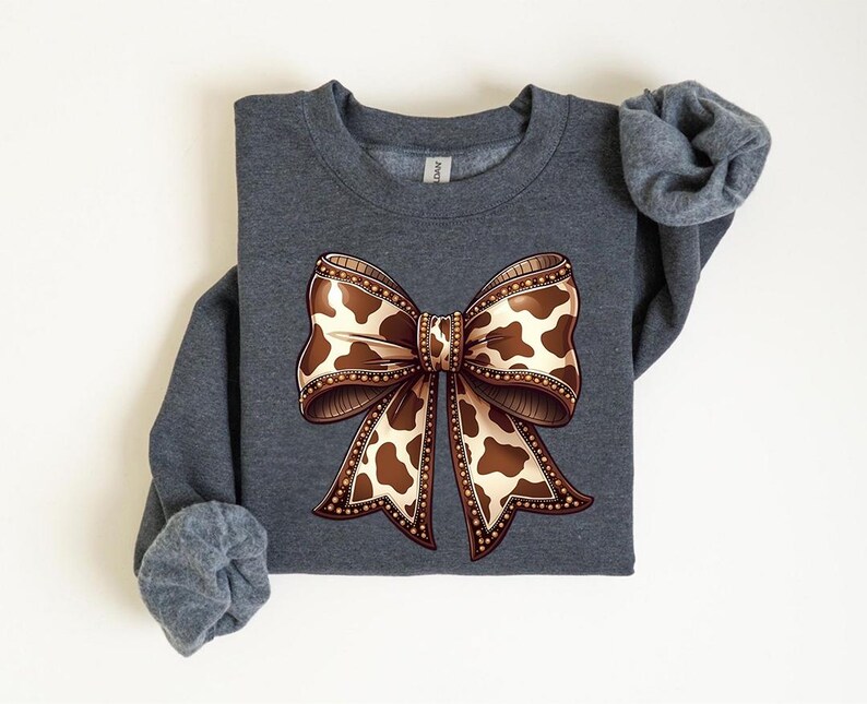May include: A dark gray sweatshirt featuring a large bow design. The bow has a brown and white cow print pattern with a decorative border. The sleeves are rolled up.