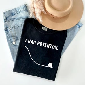 Puede incluir: Una camiseta negra de manga corta con el texto blanco I HAD POTENTIAL sobre un gráfico blanco de una bola en la parte inferior de una línea curva. La camiseta se combina con vaqueros azul claro, un sombrero beige y un collar dorado.