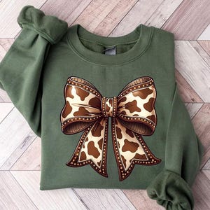May include: An olive green sweatshirt featuring a large bow design. The bow has a brown and cream cow print pattern with a decorative border. The sweatshirt has long sleeves with a ribbed cuff.