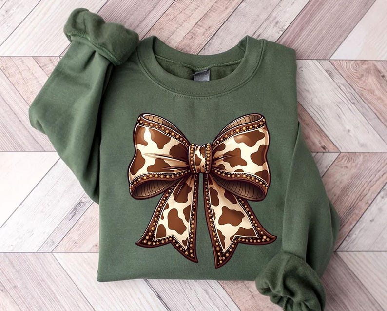 May include: An olive green sweatshirt featuring a large bow design. The bow has a brown and cream cow print pattern with a decorative border. The sweatshirt has long sleeves with a ribbed cuff.