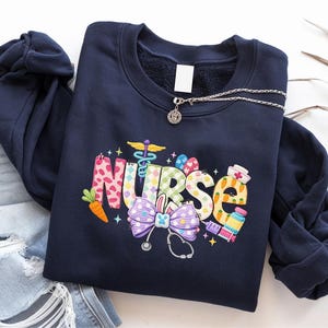 May include: A navy blue sweatshirt featuring the word "NURSE" in colourful, embroidered letters. The design includes a carrot, Easter eggs, a stethoscope, and a bow. A silver necklace with a pendant is draped across the sweatshirt.