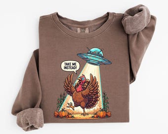 May include: Brown sweatshirt with a graphic of a turkey being beamed up by a UFO. The turkey has its wings spread and the text "TAKE ME INSTEAD!" is in a speech bubble. Pumpkins and corn stalks are at the bottom of the design.