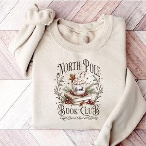A festive T-shirt featuring a cozy Christmas-themed design that reads “North Pole Book Club Hot Cocoa Served Daily.” The artwork shows a cup of hot cocoa topped with whipped cream, a candy cane, and a gingerbread cookie resting on stacked books, surrounded by holly berries and pine branches; a perfect holiday shirt for book lovers and cozy winter readers who adore festive hot chocolate and Christmas vibes.