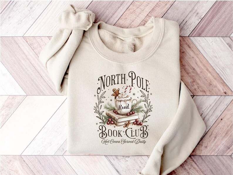 A festive T-shirt featuring a cozy Christmas-themed design that reads “North Pole Book Club Hot Cocoa Served Daily.” The artwork shows a cup of hot cocoa topped with whipped cream, a candy cane, and a gingerbread cookie resting on stacked books, surrounded by holly berries and pine branches; a perfect holiday shirt for book lovers and cozy winter readers who adore festive hot chocolate and Christmas vibes.