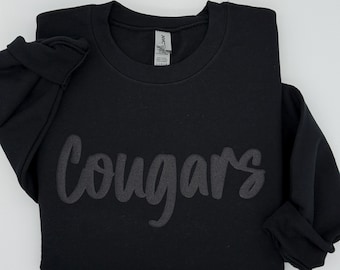May include: Black crew neck sweatshirt with the word "Cougars" in a darker tone across the chest. The sleeves are folded up. The sweatshirt is on a white background.