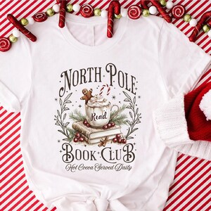 A festive T-shirt featuring a cozy Christmas-themed design that reads “North Pole Book Club Hot Cocoa Served Daily.” The artwork shows a cup of hot cocoa topped with whipped cream, a candy cane, and a gingerbread cookie resting on stacked books, surrounded by holly berries and pine branches; a perfect holiday shirt for book lovers and cozy winter readers who adore festive hot chocolate and Christmas vibes.